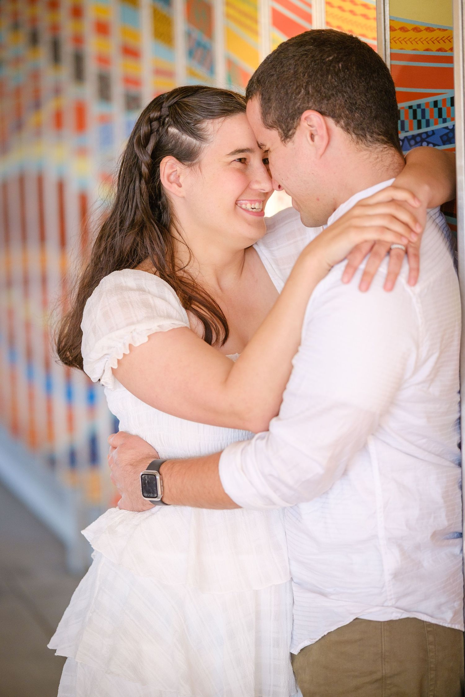 Smiling couple in white outfits embracing tenderly in front of a colorful mosaic wall.