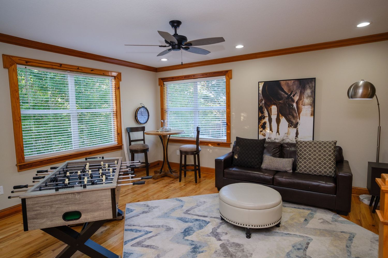 Modern living room with brown leather couch, foosball table, wood trim windows and blue patterned area rug on hardwood floors.