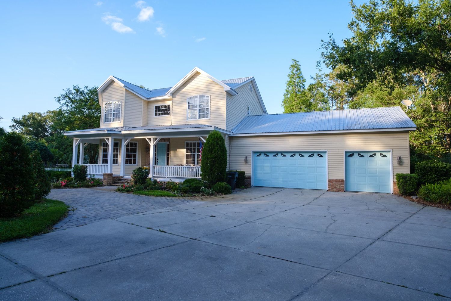 Large white two-story suburban house with wrap-around porch and three-car garage on expansive concrete driveway.