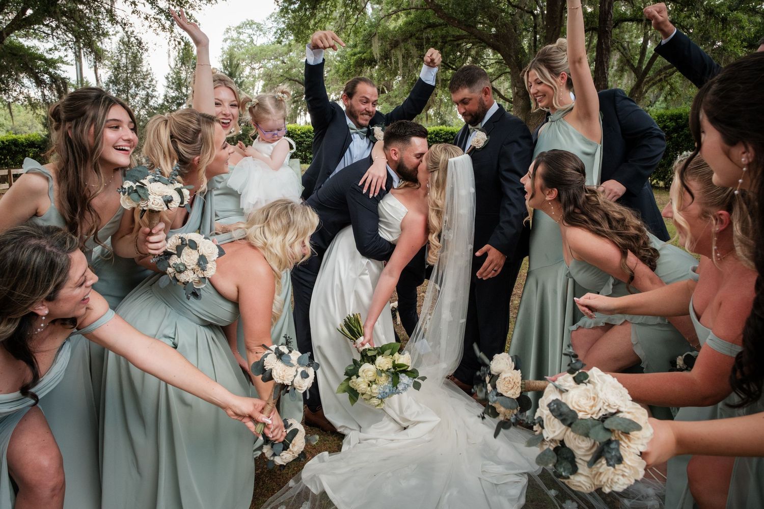 Wedding party celebrates bride and groom kissing, bridesmaids in sage green dresses holding white bouquets outdoors.
