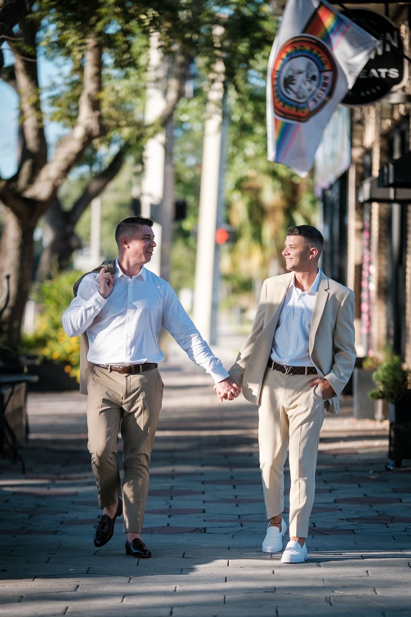 Two men in suits hold hands while walking on a sunny tree-lined sidewalk with a pride flag in the background.