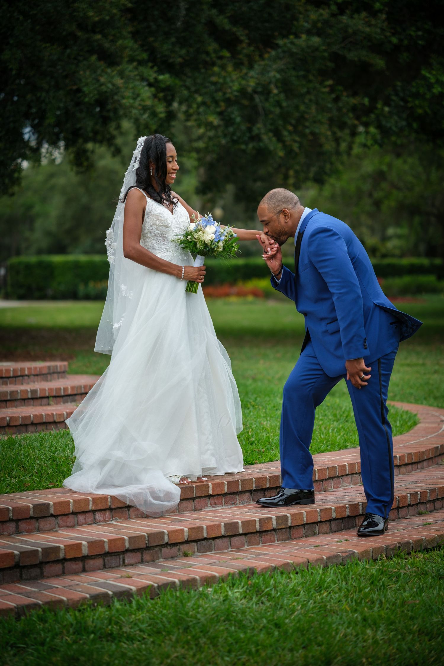 Groom in blue suit kisses bride's hand on brick steps outdoors during wedding ceremony.