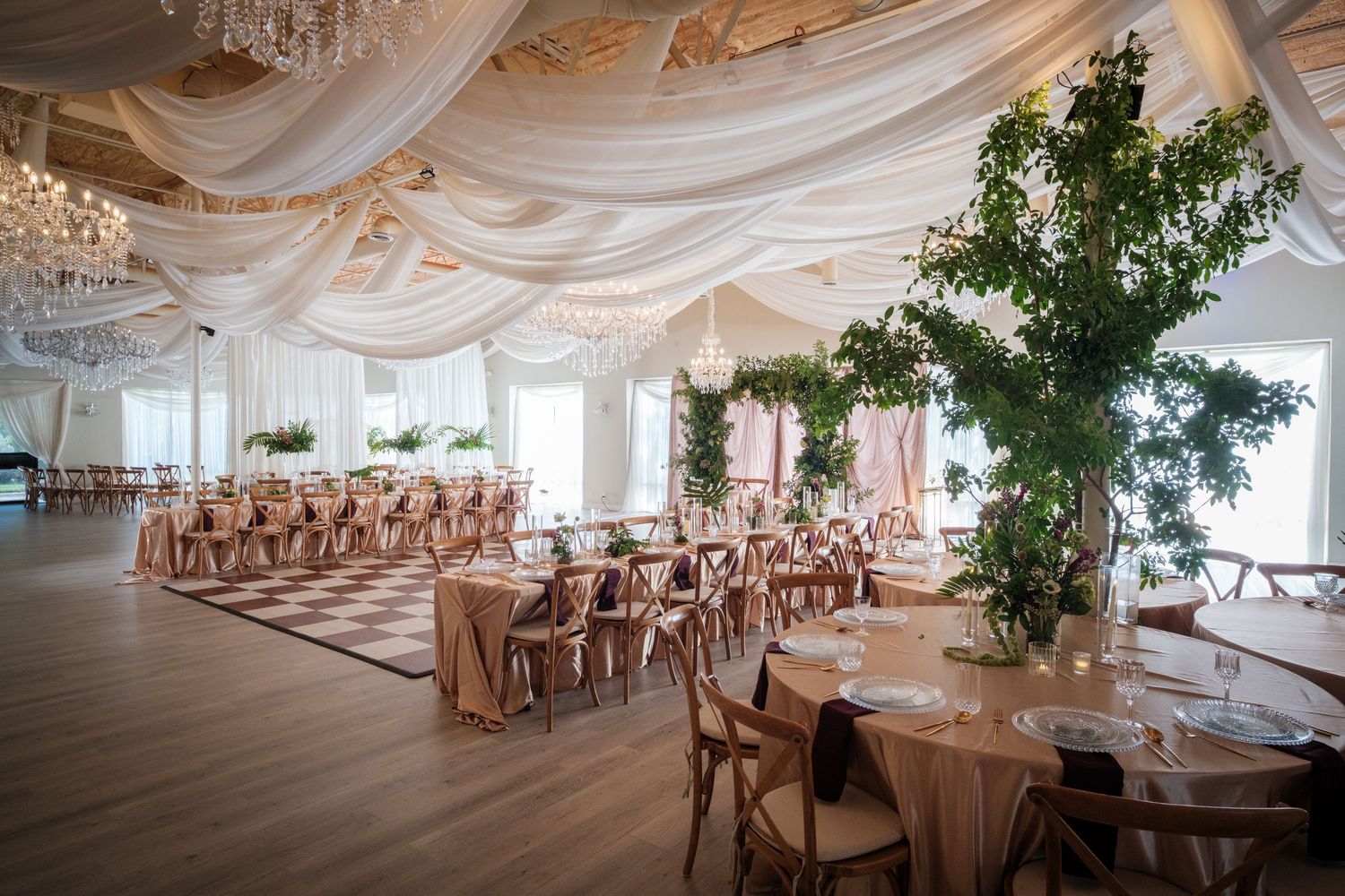 Elegant wedding reception hall with white draped ceiling, chandeliers, wooden chairs, and lush greenery centerpieces.