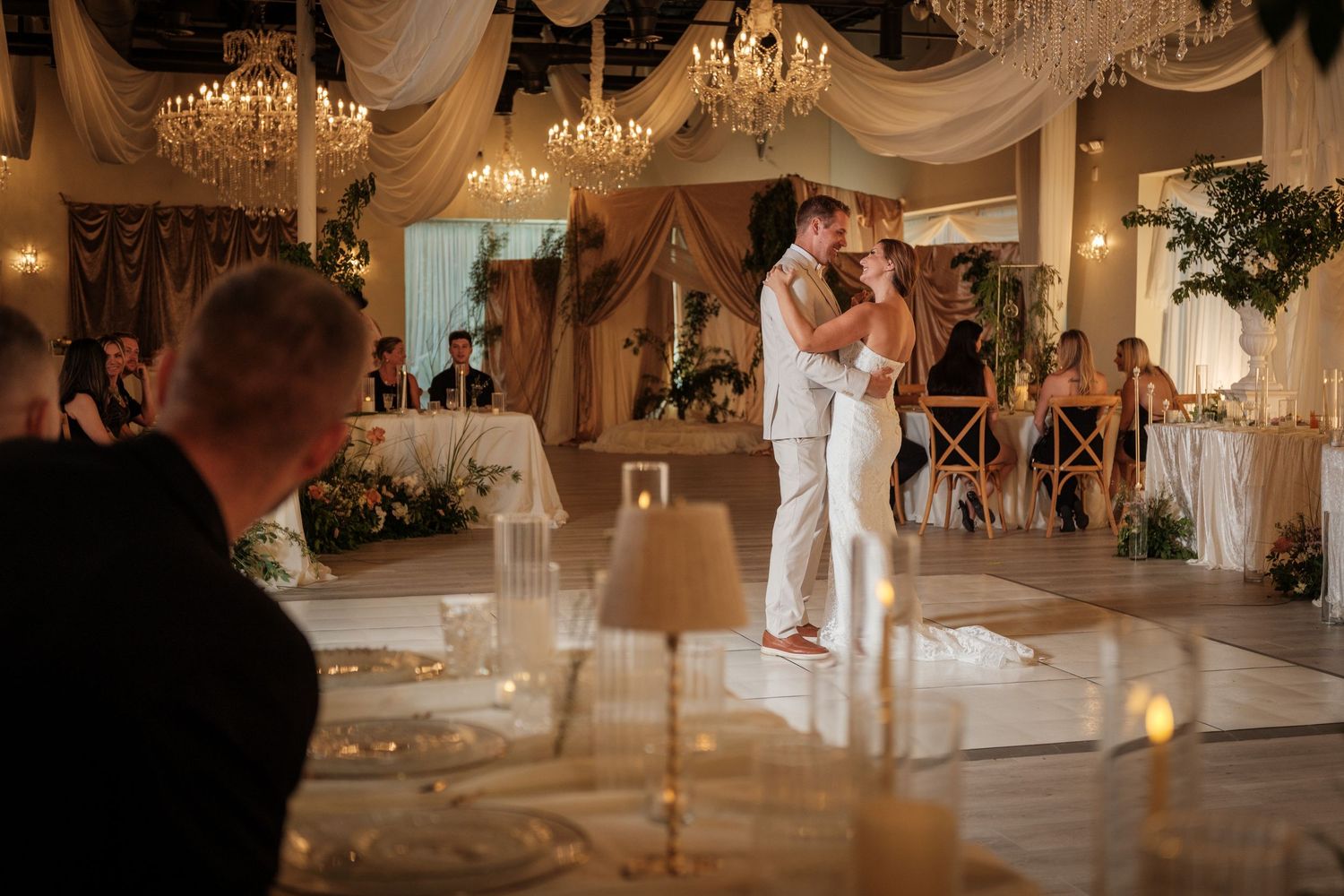 Couple sharing first dance at elegant wedding reception with chandeliers, draped ceilings, and candlelit tables.