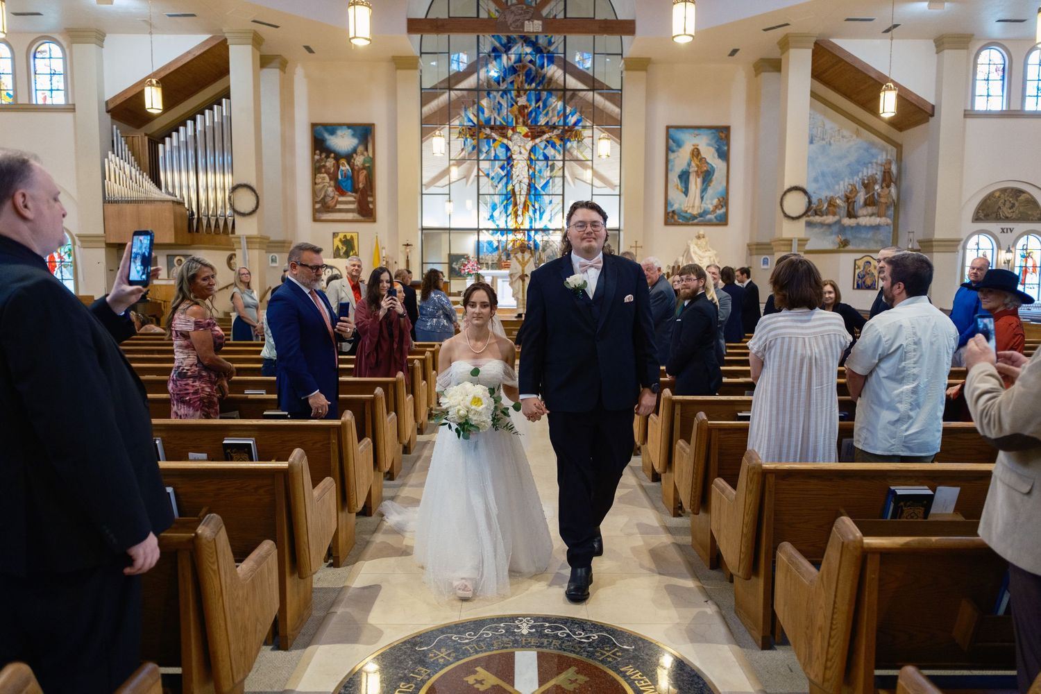 Bride and groom walking down church aisle after wedding ceremony, surrounded by guests in wooden pews.