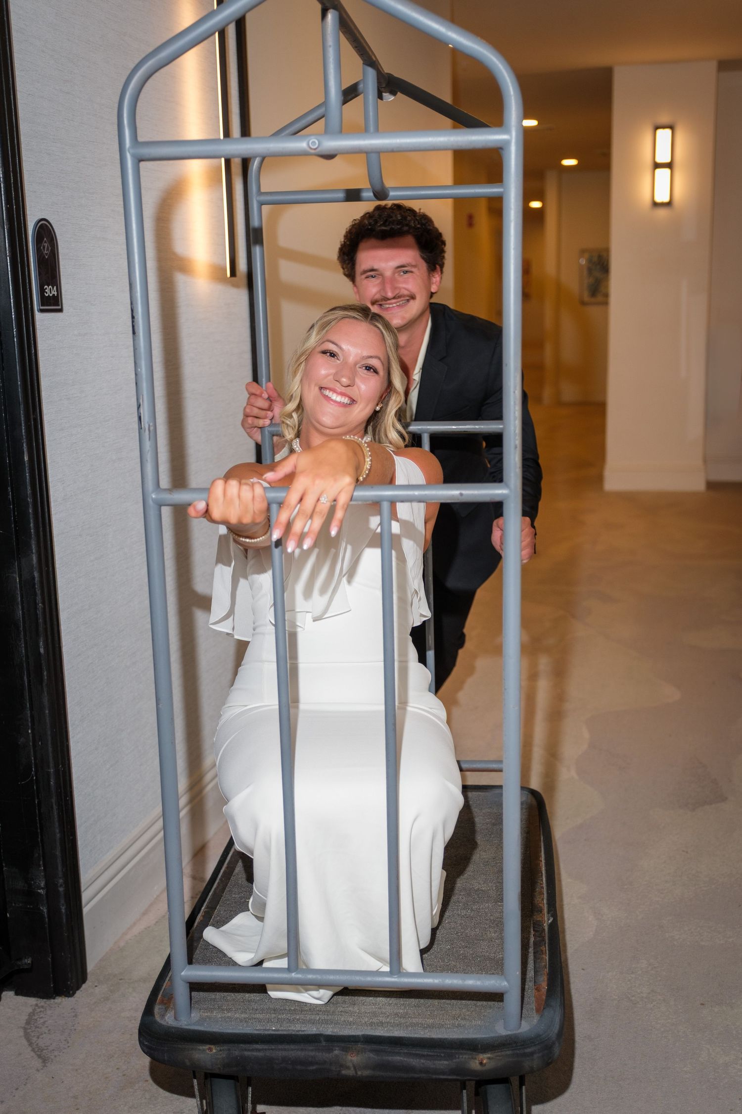Newlyweds pose playfully on a hotel luggage cart, bride in white dress showing ring, groom in dark suit smiling behind her.