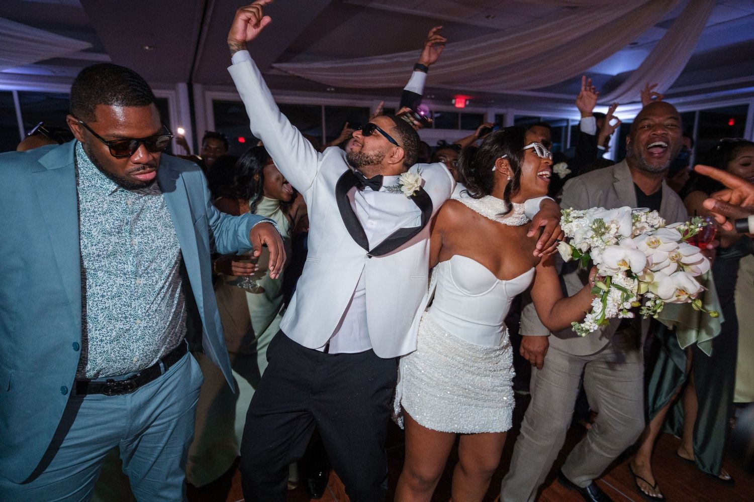 Joyful wedding reception dance floor with bride in white dress and groom in tuxedo celebrating with guests under tent lighting.