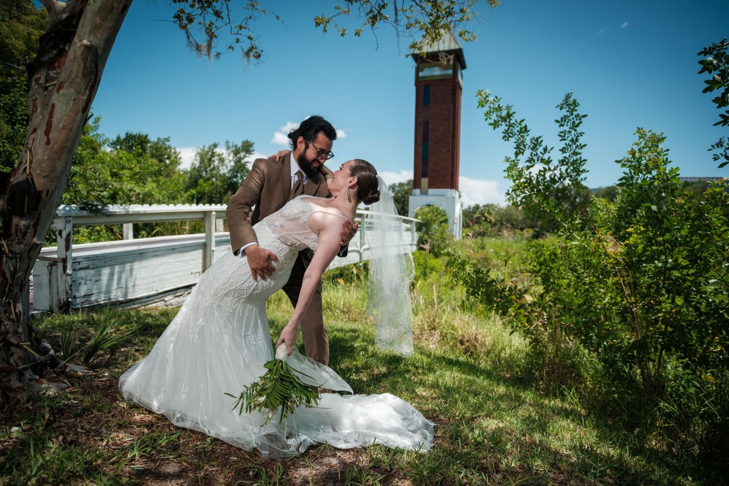 Bride and groom share a romantic dip kiss outdoors under a tree with a clock tower in the background on their wedding day.