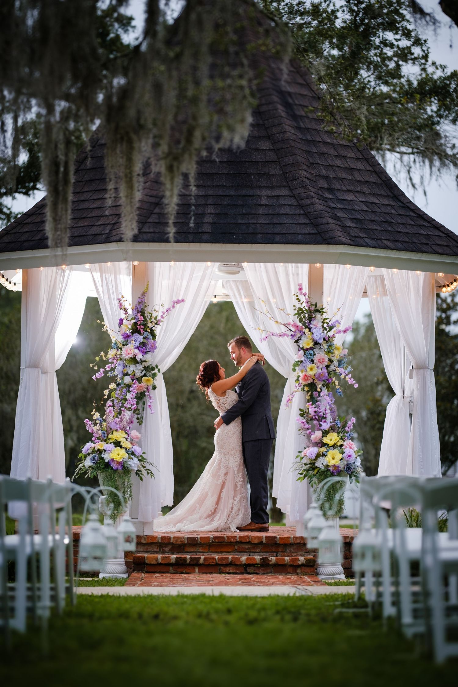 A couple shares their first kiss under a white gazebo decorated with purple and white flowers at an outdoor wedding ceremony.