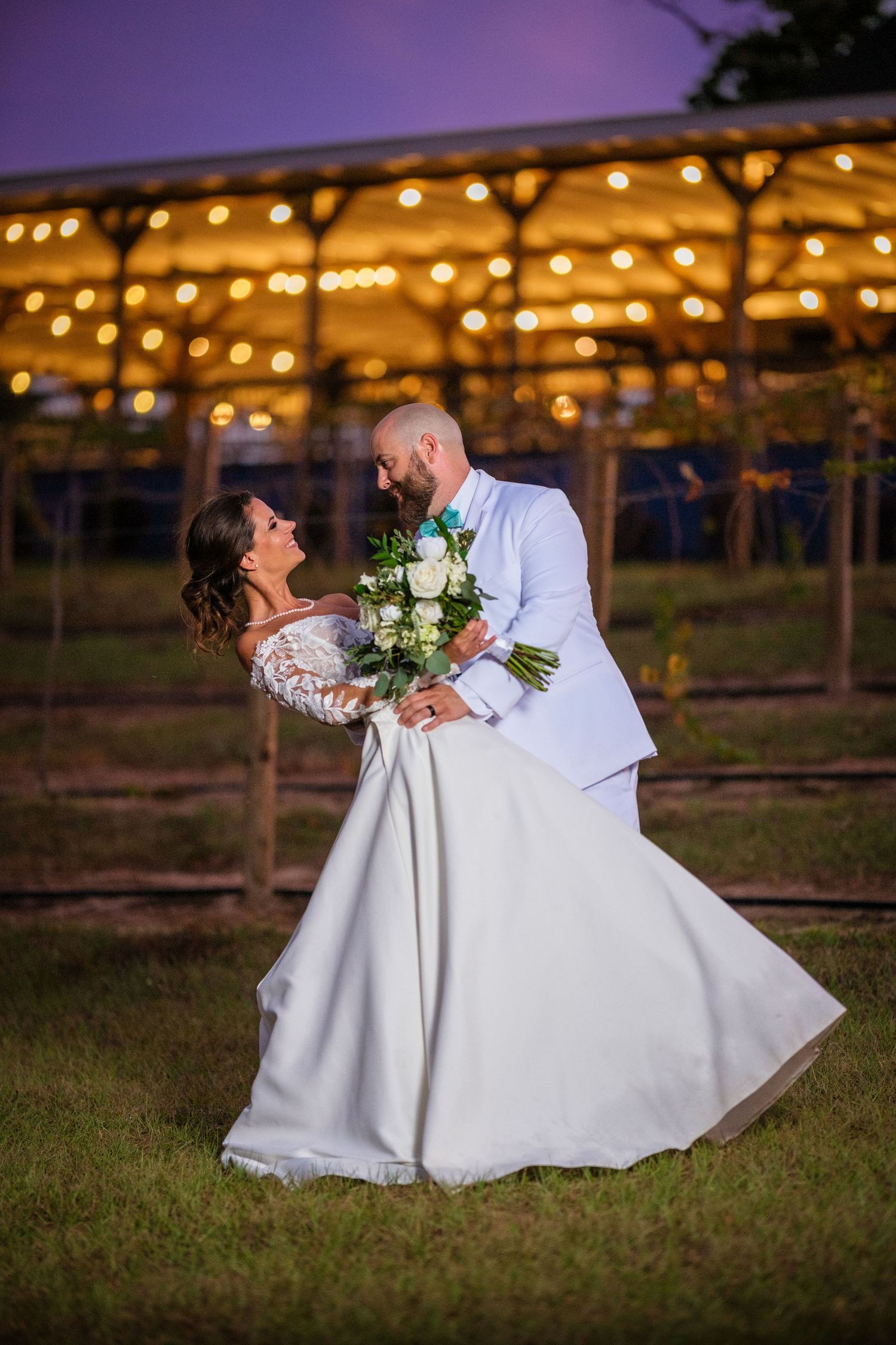 Romantic wedding couple dips and kisses at sunset under twinkling string lights in an outdoor venue.
