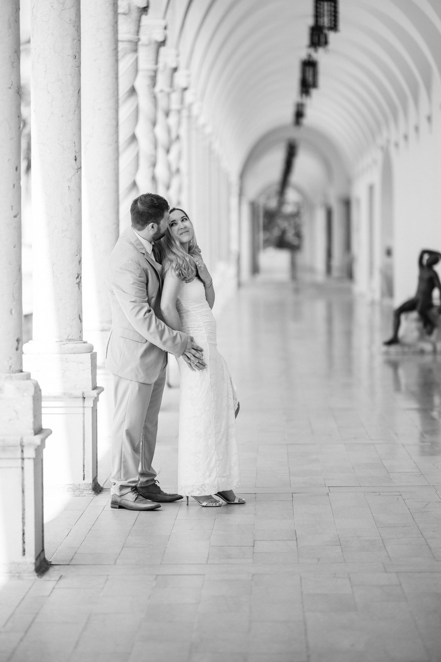 Black and white photo of a couple embracing in an elegant columned hallway, woman in white dress, man in suit.