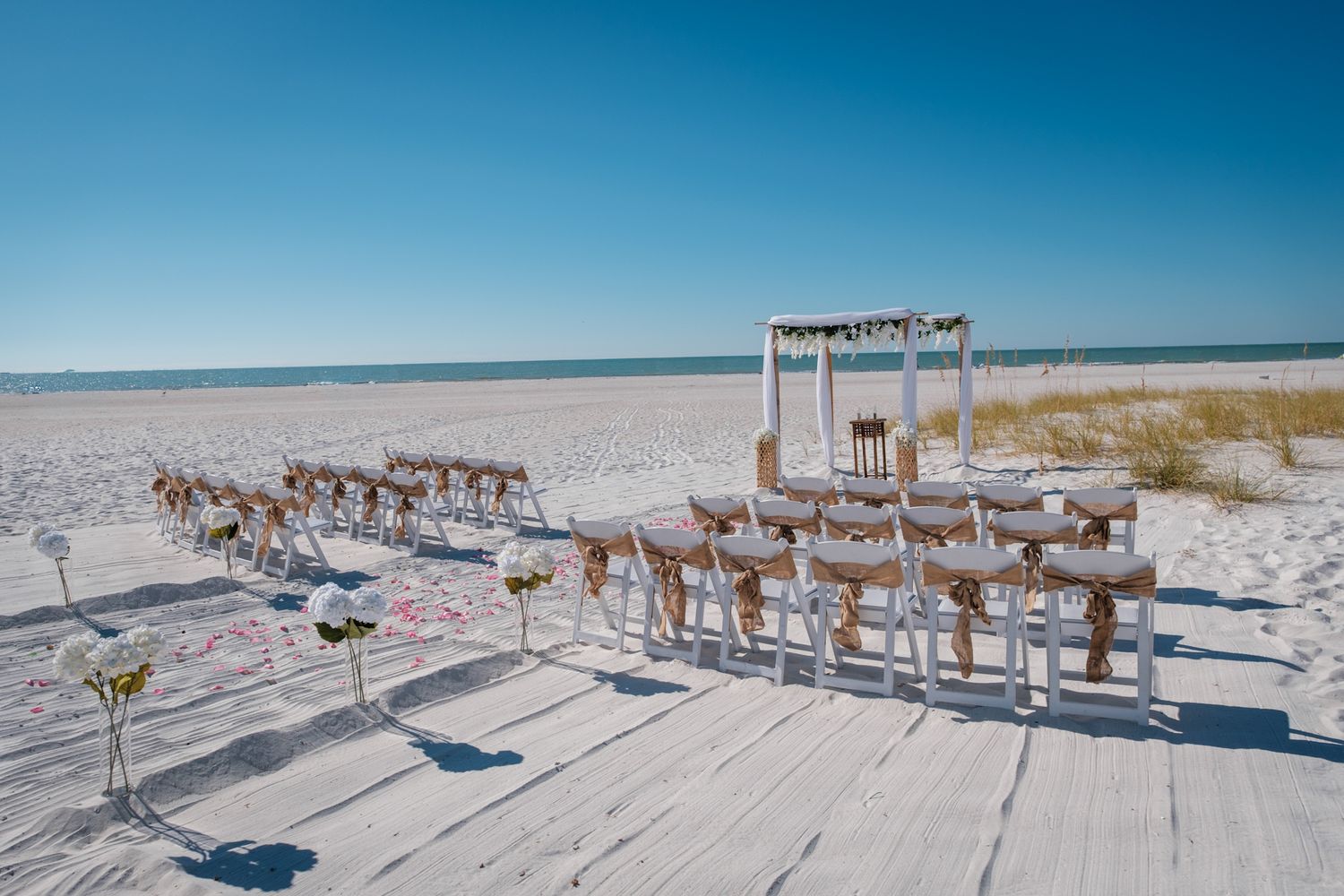 White wooden chairs arranged for a beach wedding ceremony with an arch and ocean backdrop on a sunny day.