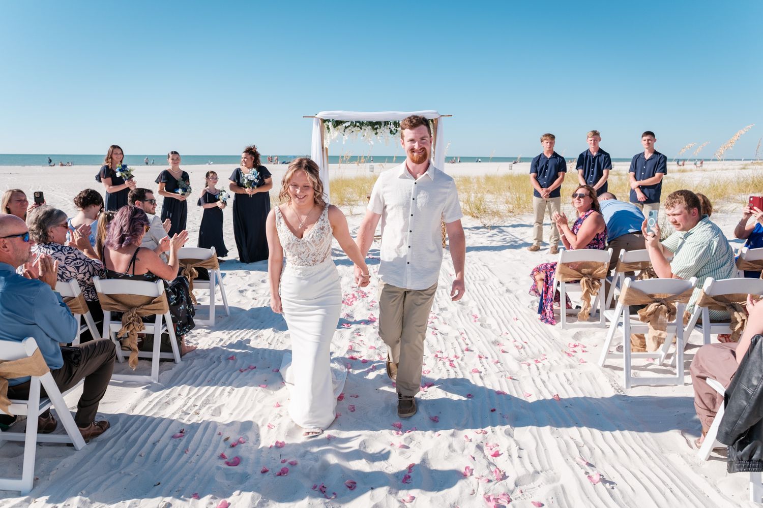 Wedding guests seated in white chairs on a sandy beach with ocean views during an outdoor ceremony.