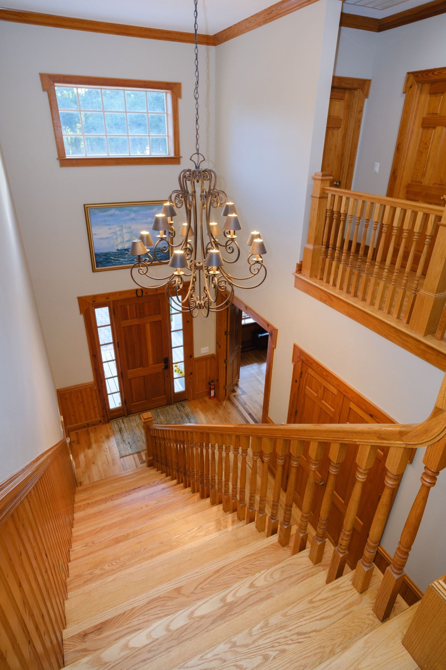 Elegant wooden staircase with oak railings and hardwood steps descending beneath a grand chandelier in a sunlit foyer.