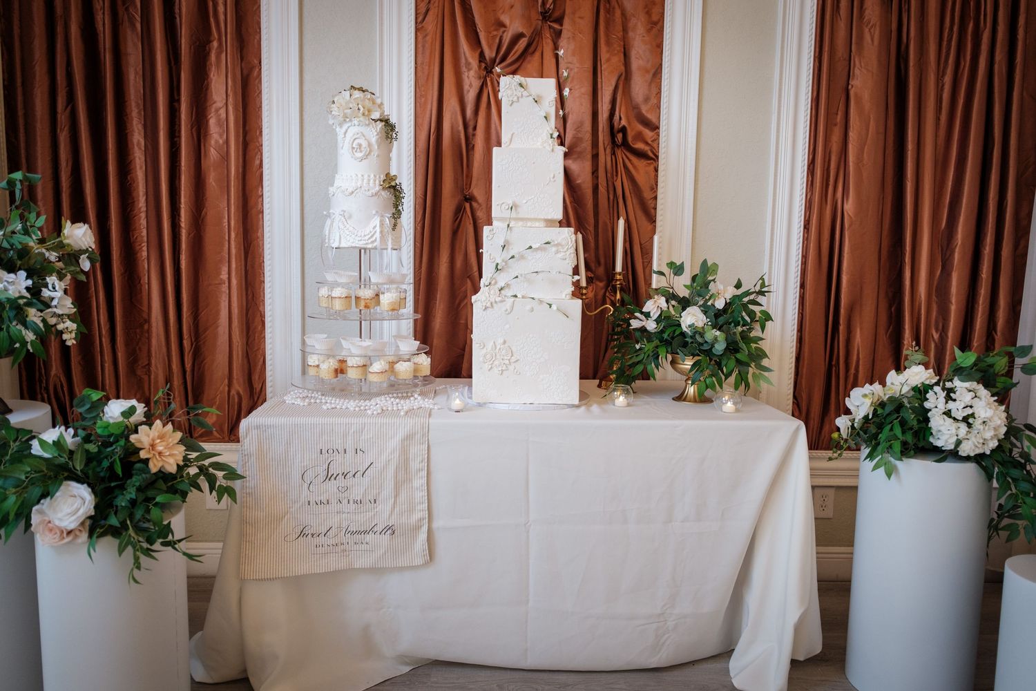 Elegant wedding cake display on white draped table with floral arrangements and copper curtain backdrop.