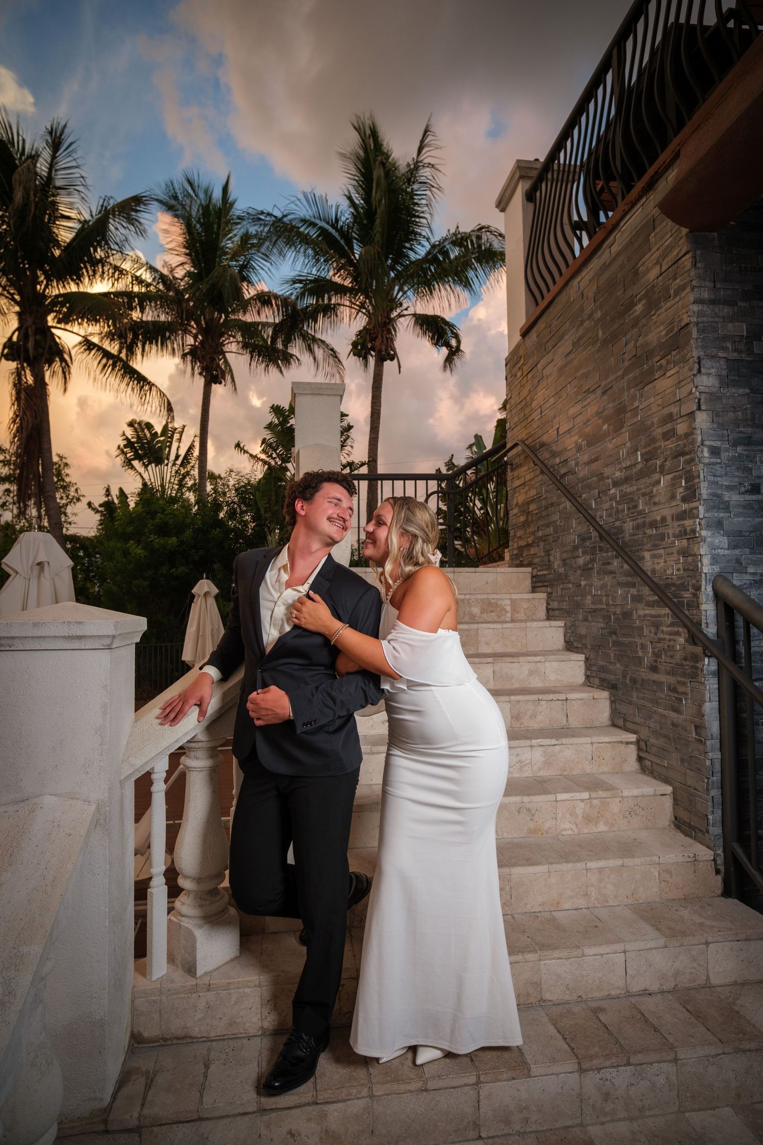 A couple in wedding attire shares a romantic moment on marble stairs with palm trees and sunset sky backdrop.