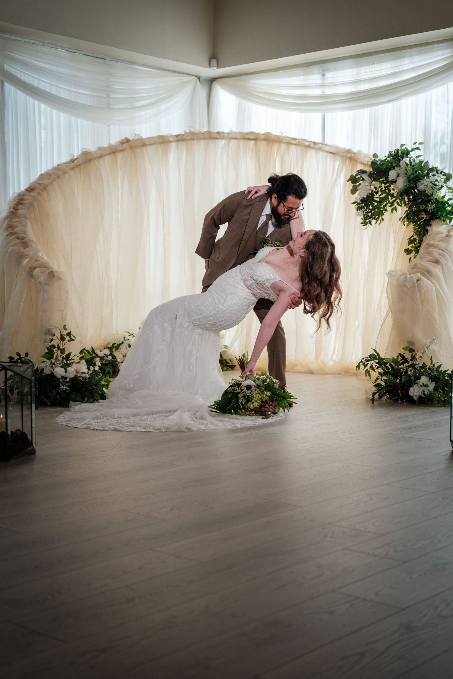 Groom dips bride for a romantic kiss in front of an elegant floral arch backdrop at their wedding reception.