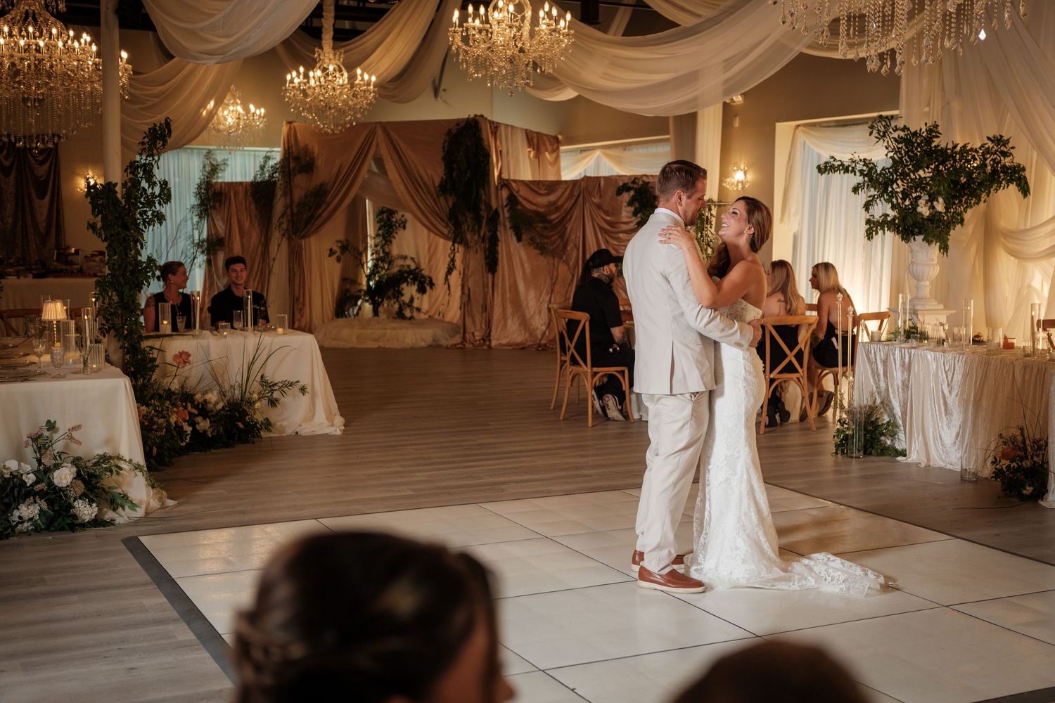 Bride and groom share their first dance on a white dance floor in an elegant wedding reception venue with chandeliers.