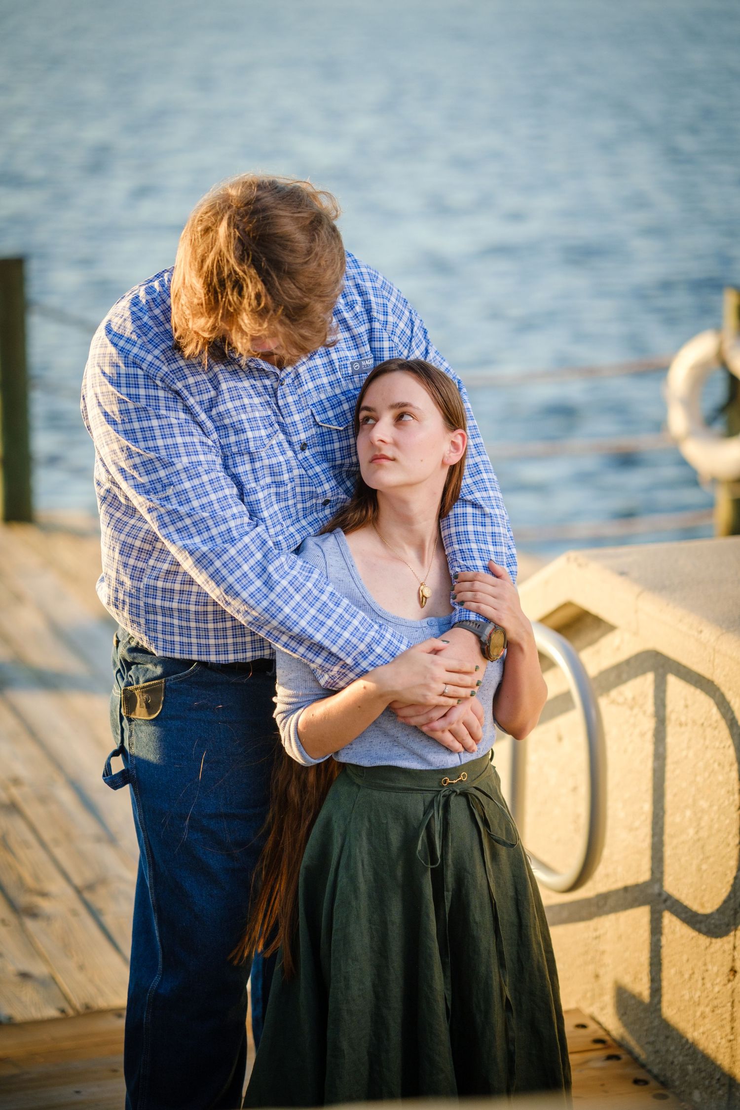 Couple embracing on a waterfront dock at sunset, woman in green skirt looks up at man in plaid shirt.