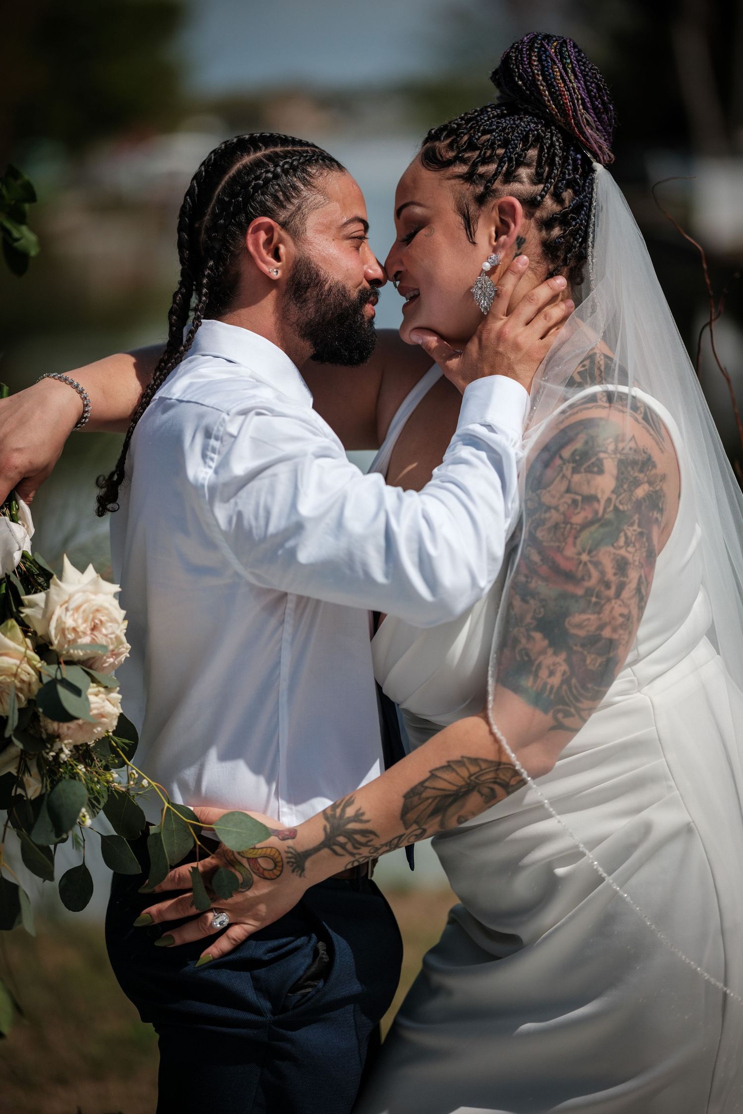 A bride and groom share a romantic almost-kiss moment outdoors on their wedding day, both wearing braids and elegant attire.