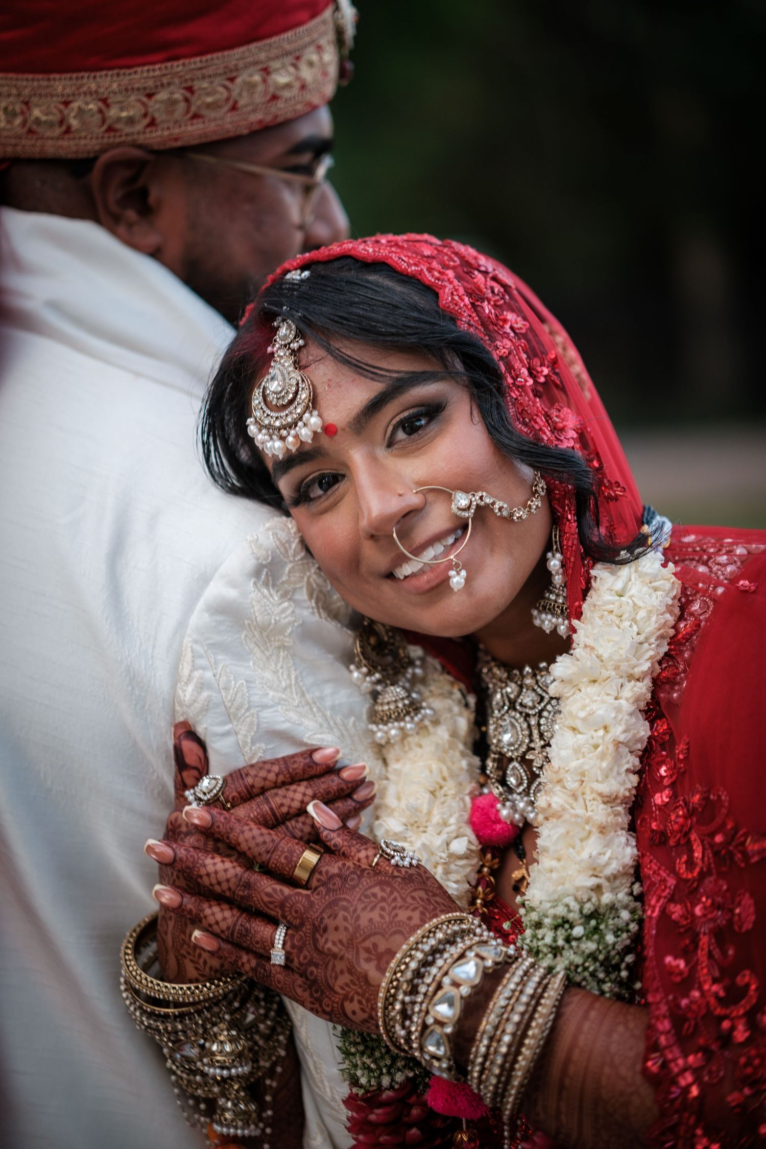 Indian bride in red saree with mehndi hands smiles leaning on groom during traditional Hindu wedding ceremony.