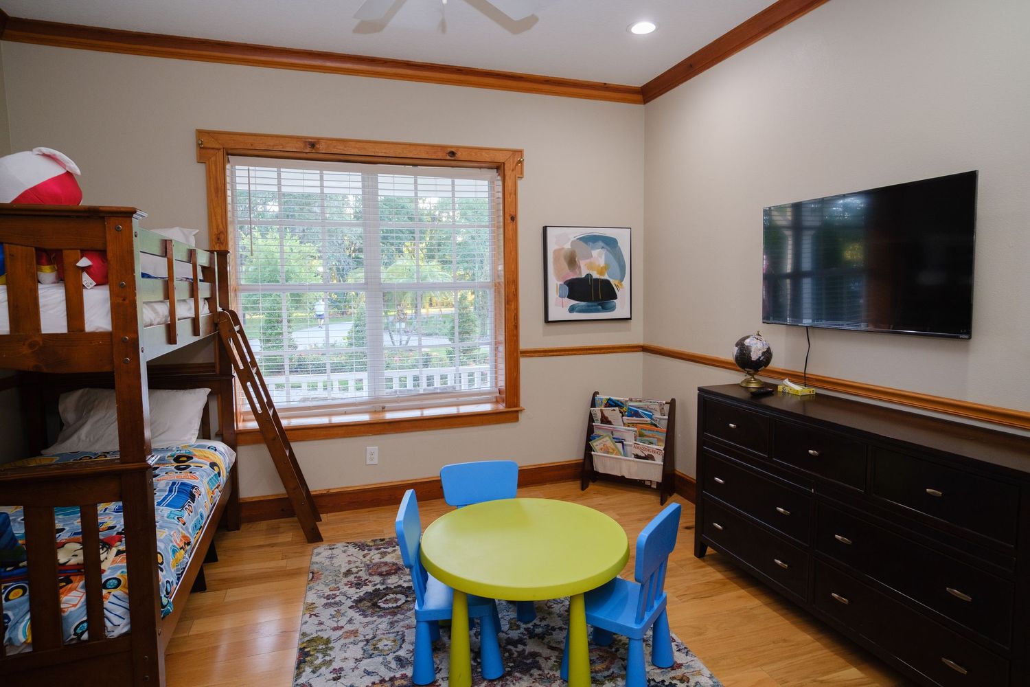 A child's playroom features a bunk bed, colorful round table, black dresser, and wall-mounted TV with natural light from windows.