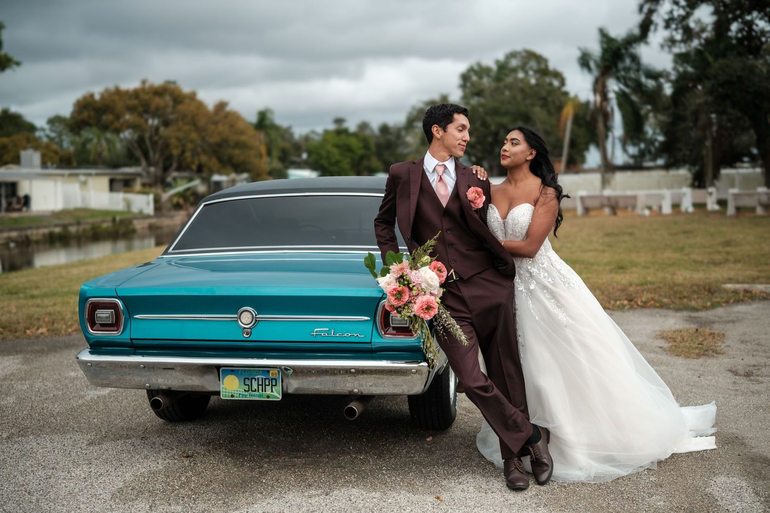 A wedding couple poses with a vintage turquoise car on a cloudy autumn day.