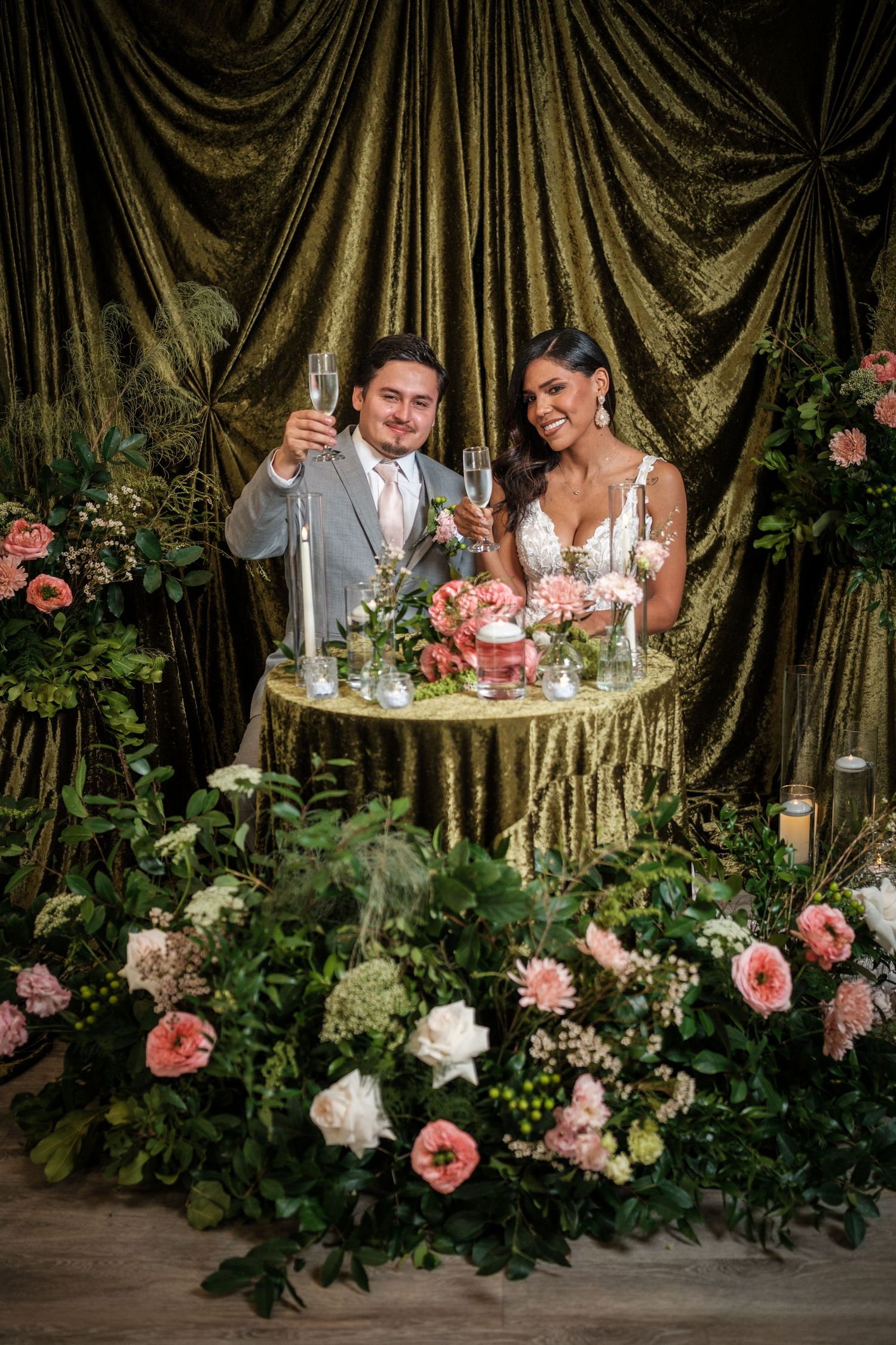 A romantic wedding scene with a couple toasting at an elegant table adorned with pink roses and gold draped backdrop.