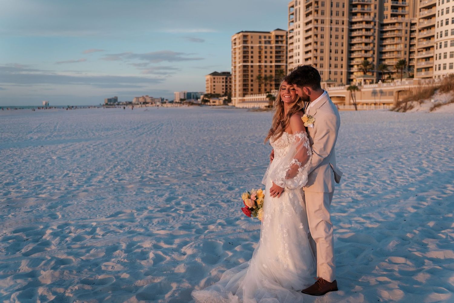 Couple in wedding attire embraces on a white sandy beach at sunset with coastal high-rise buildings in background.
