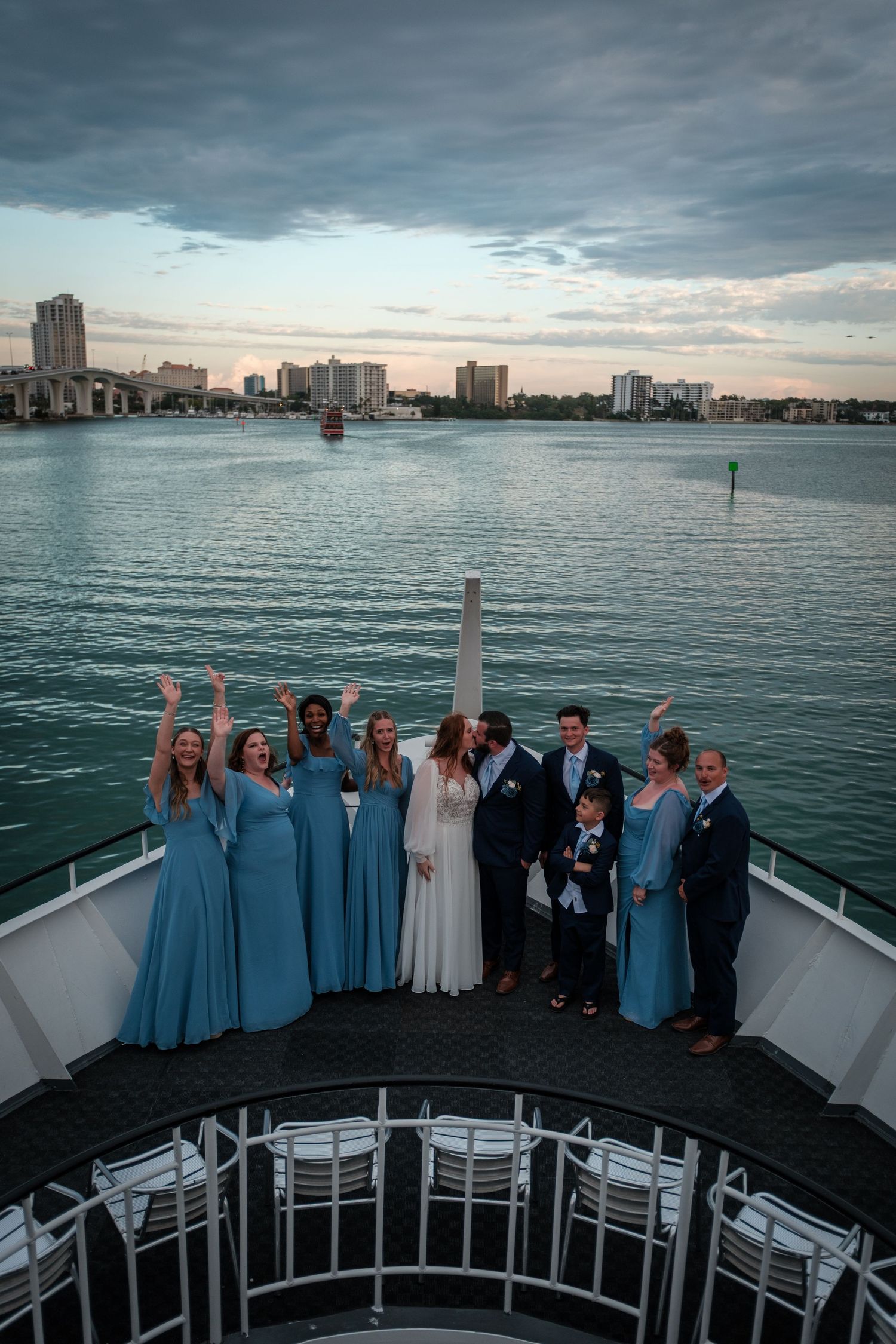 Wedding party in blue dresses celebrating on a yacht deck at sunset with a city skyline and water in the background.