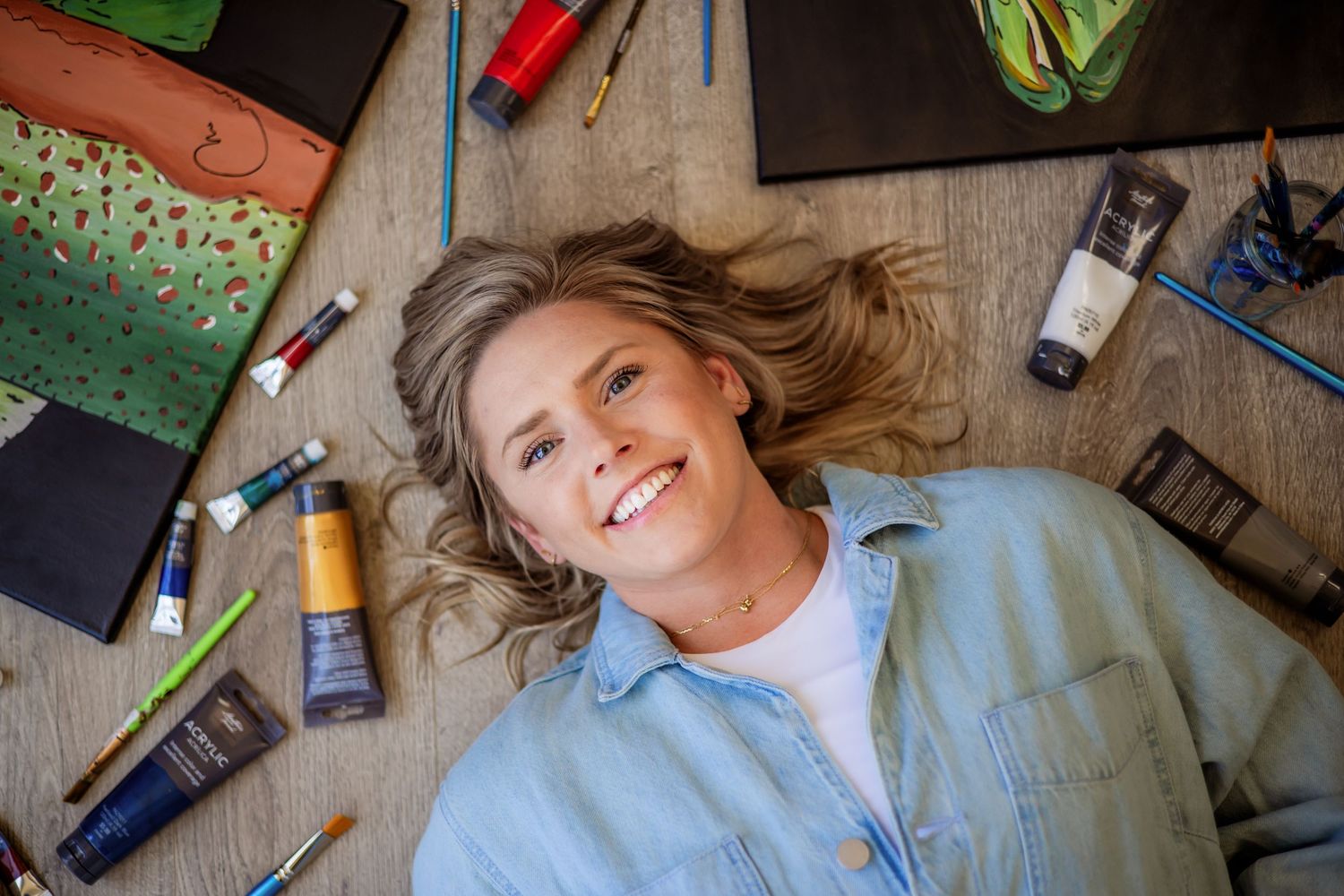 Smiling person laying on floor surrounded by art supplies and paint tubes in natural light.