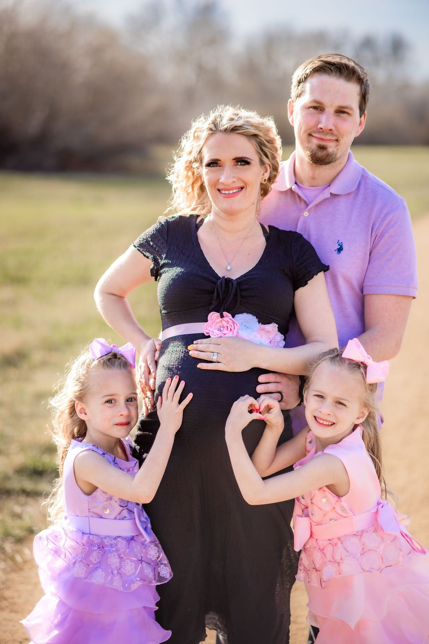 Family portrait in field with expectant mother wearing black dress and two children in purple dresses.