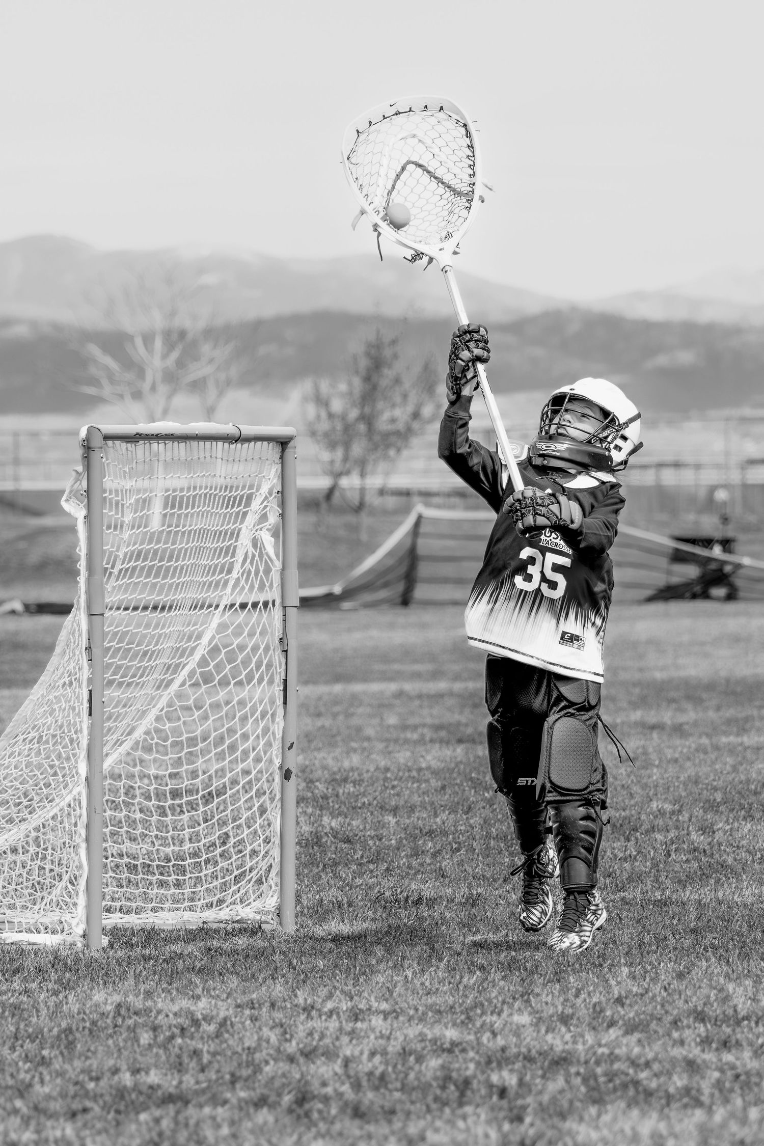 Young lacrosse player in uniform reaches high with stick near goal net in black and white photo.
