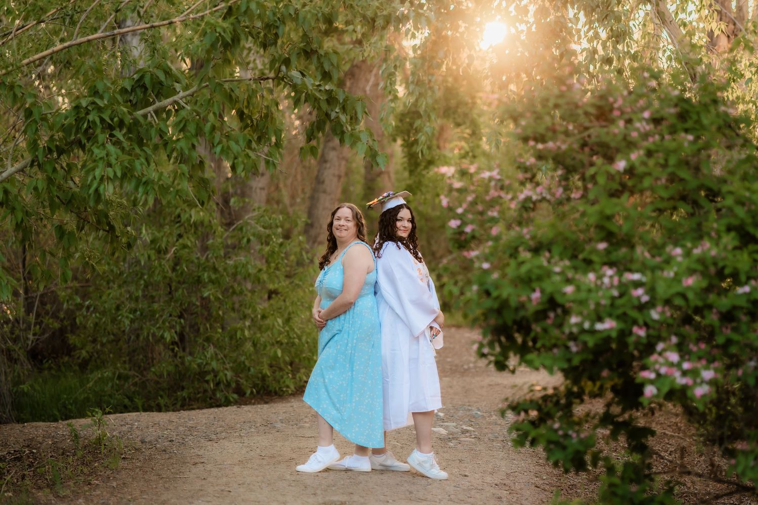 Two people in light colored dresses stand back-to-back on a nature path during golden hour sunset.