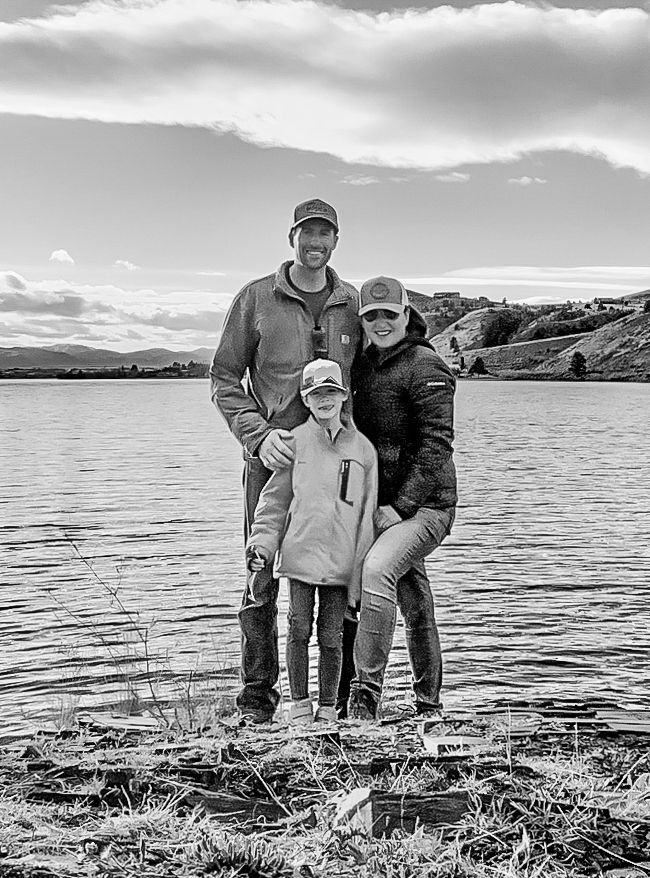 Family of three posing together by a scenic lake with mountains in the background.