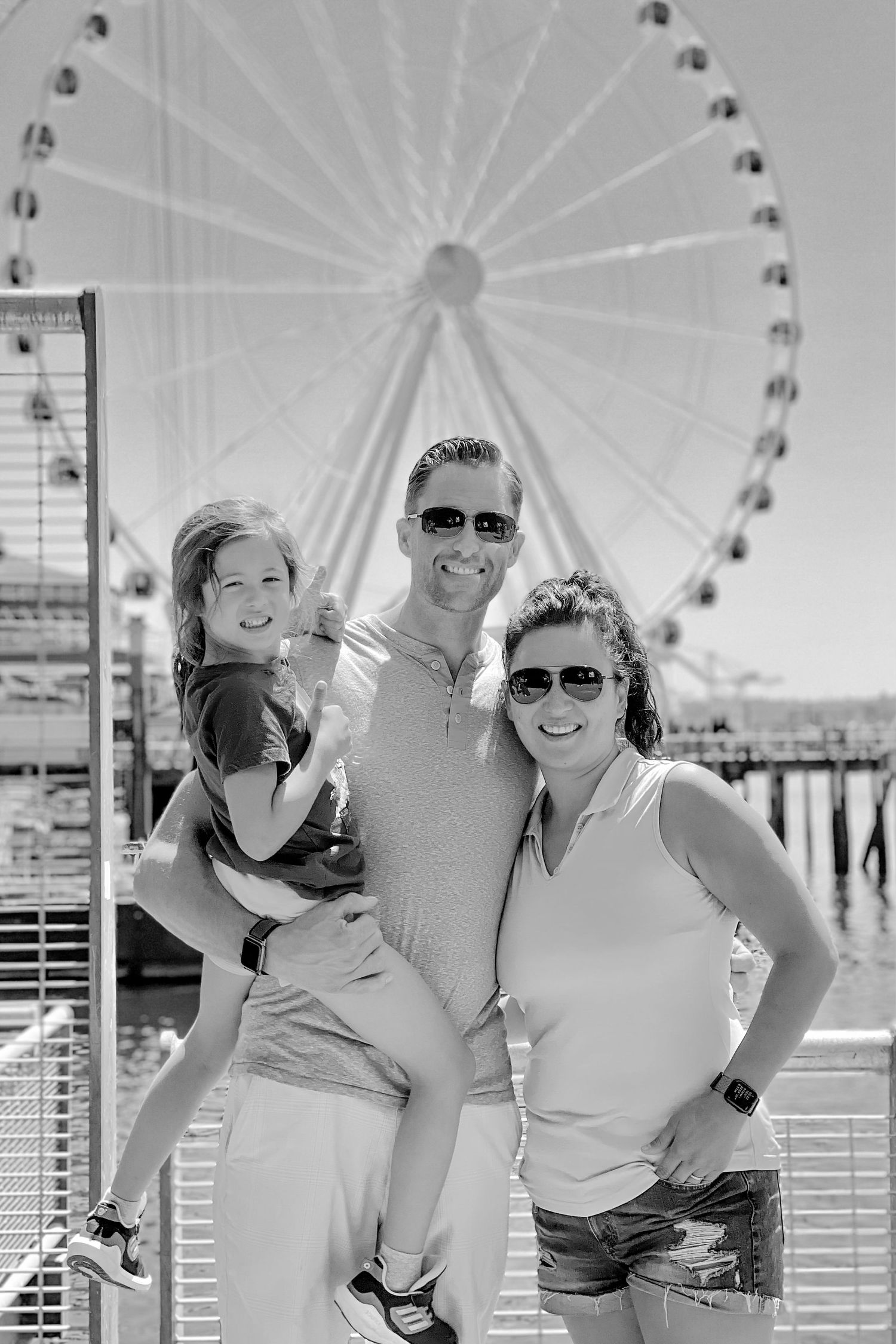 Family enjoying a sunny day together in front of a large Ferris wheel at a pier or boardwalk.