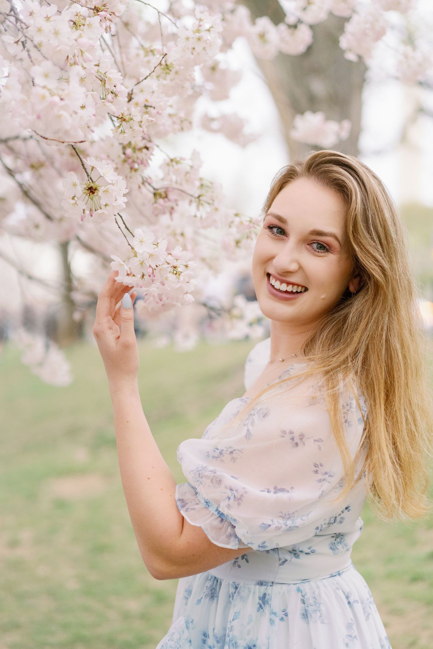 Woman in blue floral dress touches cherry blossoms while smiling on a spring day.