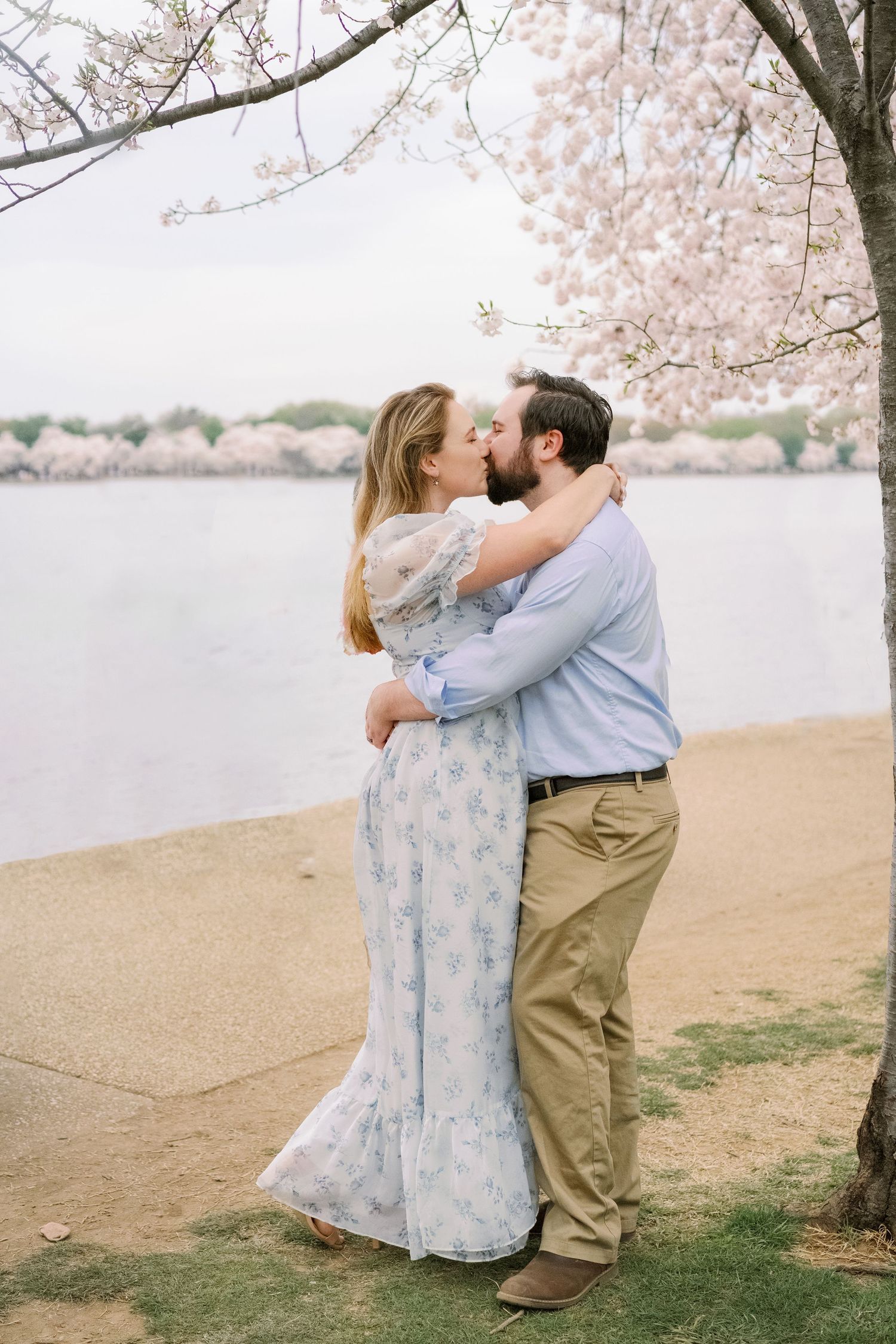 Romantic embrace under cherry blossoms with water and pink petals in background.