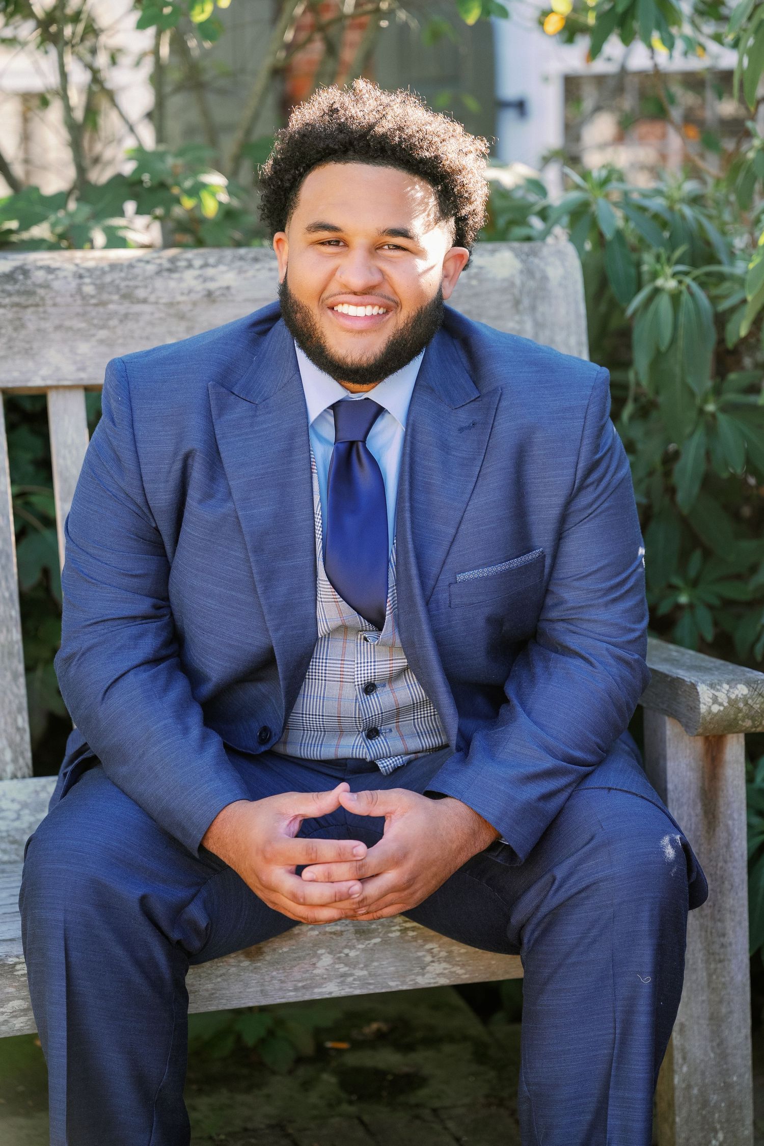 A person in a navy blue suit and vest poses casually on a wooden bench in an outdoor setting.