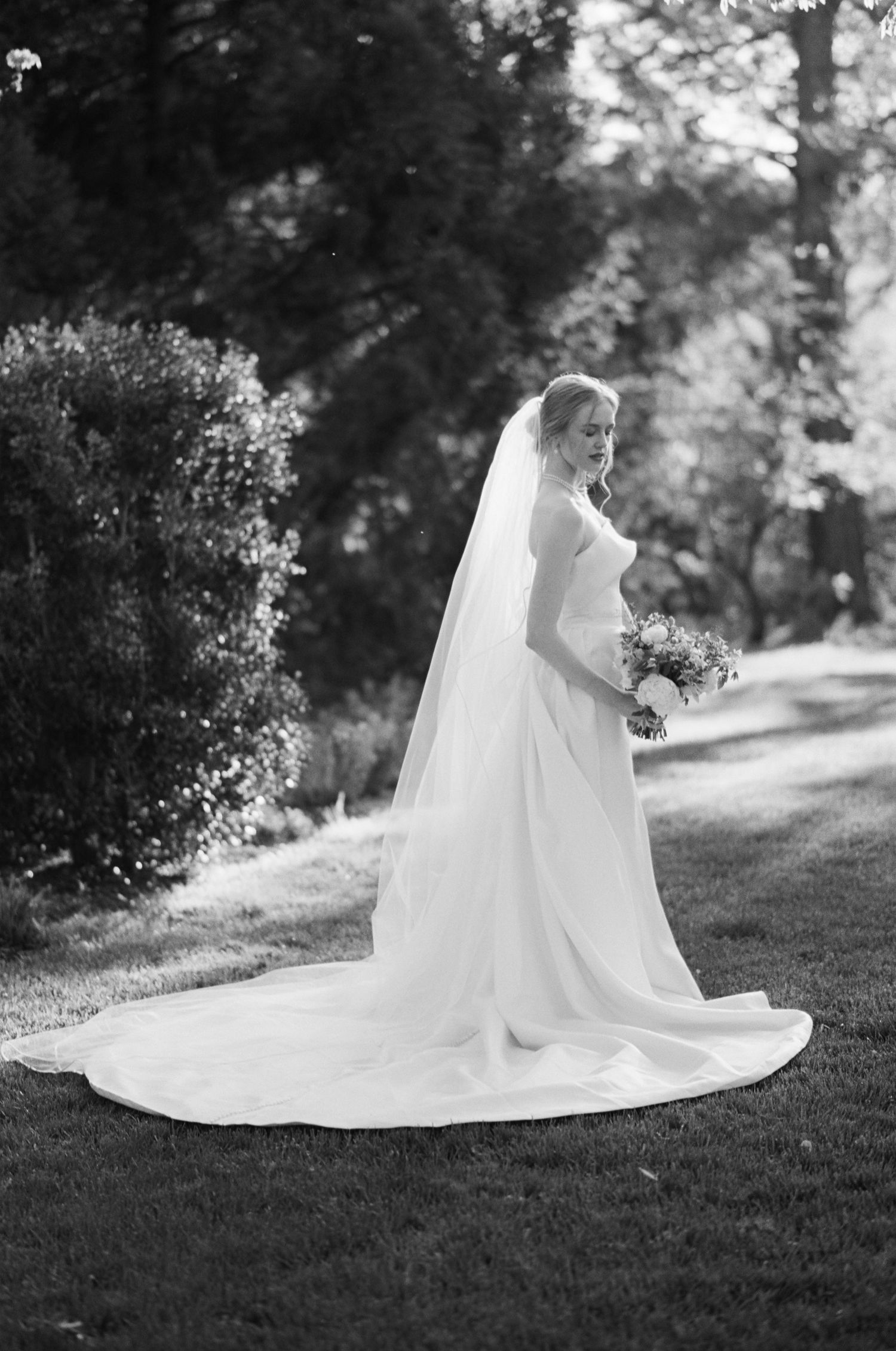 A bride in classic white gown and veil poses in a garden setting.