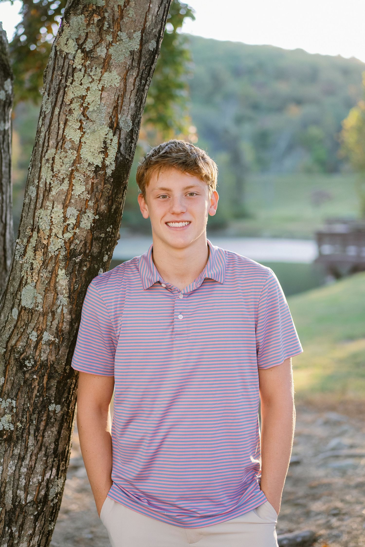 Casual outdoor portraits by tree trunk with lake and rolling hills in background.