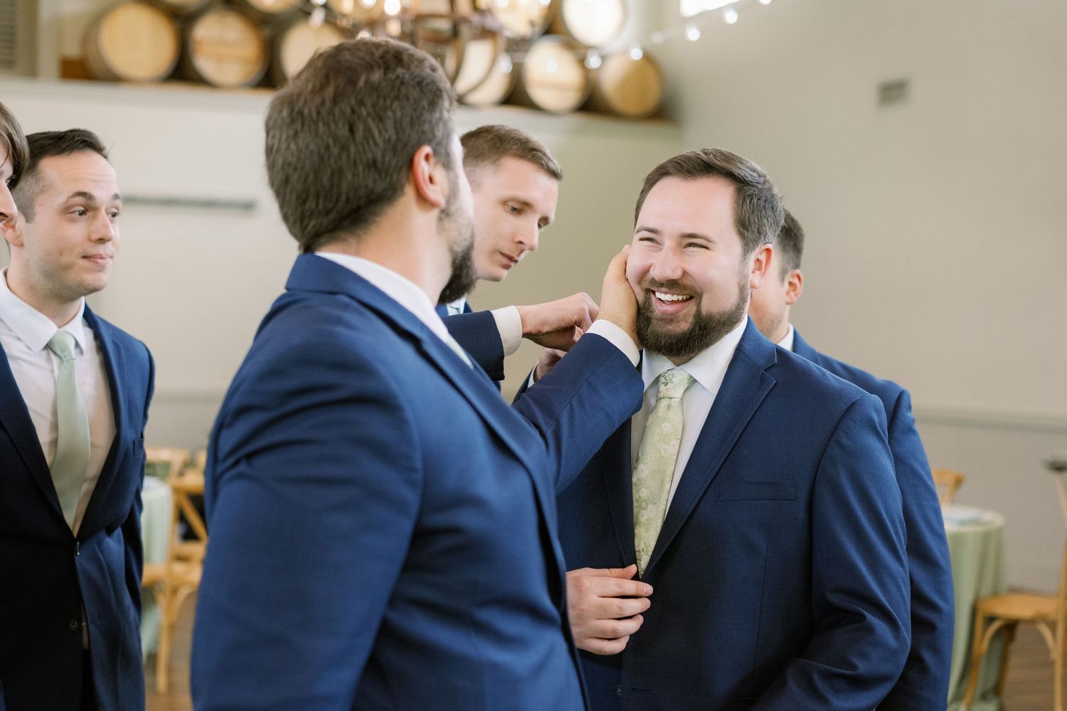 Wedding party members in navy suits share a lighthearted moment while getting ready in a venue with wooden barrels.