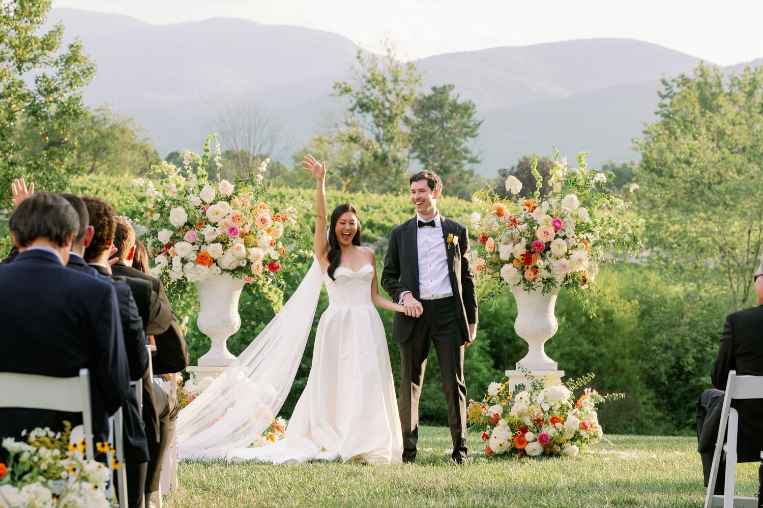 A bride and groom stand together at an outdoor wedding ceremony surrounded by floral arrangements and guests.