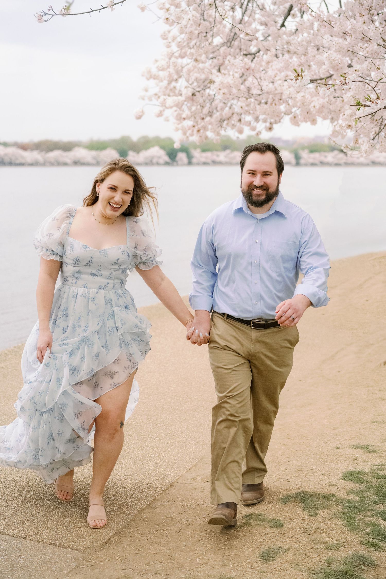 Couple walking hand in hand along cherry blossom-lined path by the water.
