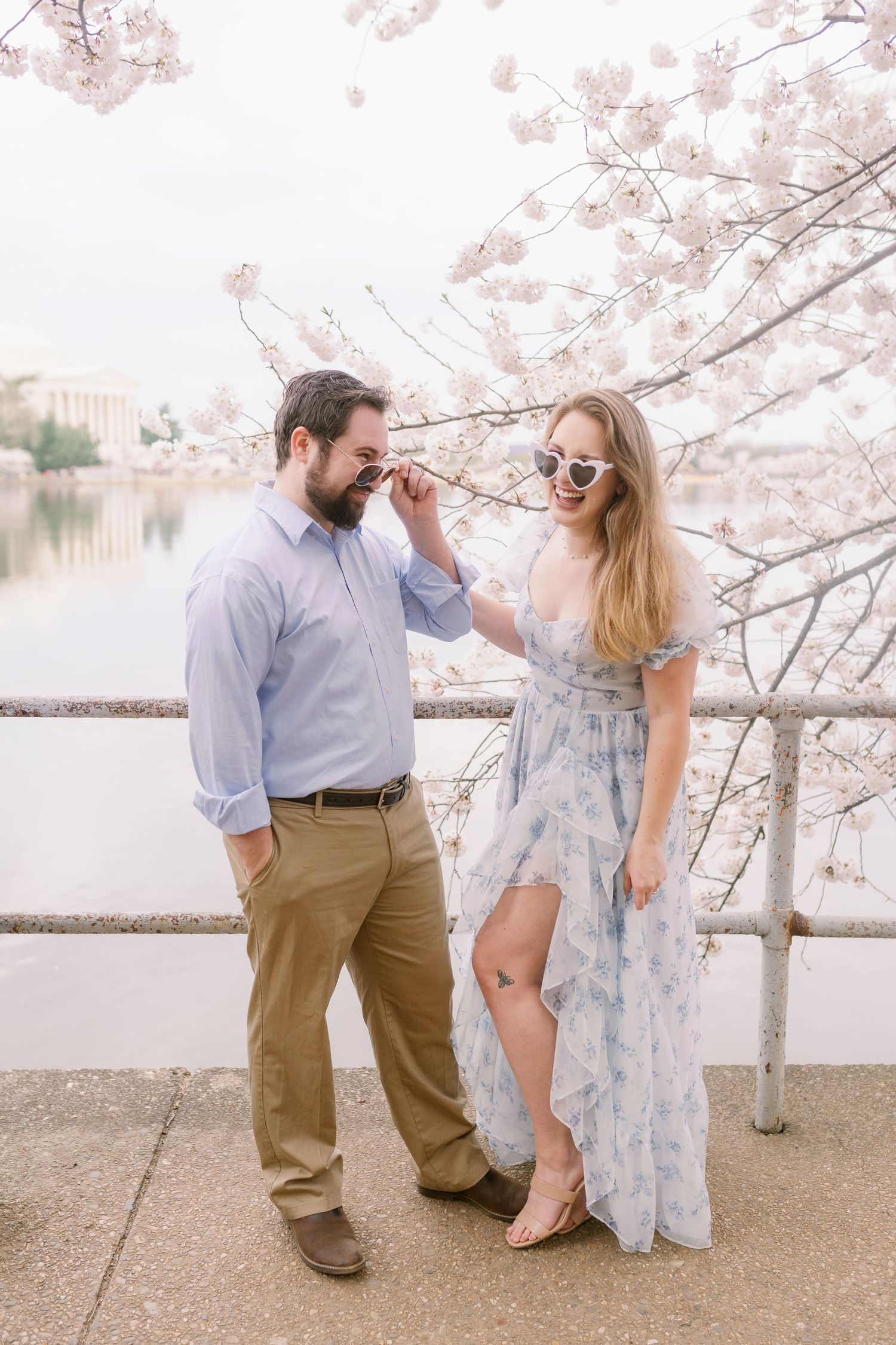 A couple shares a playful moment under blooming cherry blossoms by a waterfront railing.