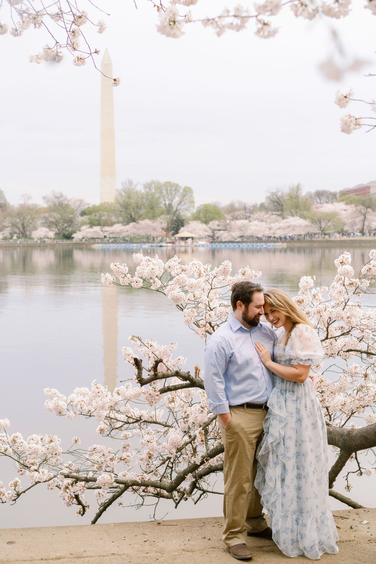 Spring cherry blossoms surround a couple by the water with the Washington Monument reflecting in the basin.