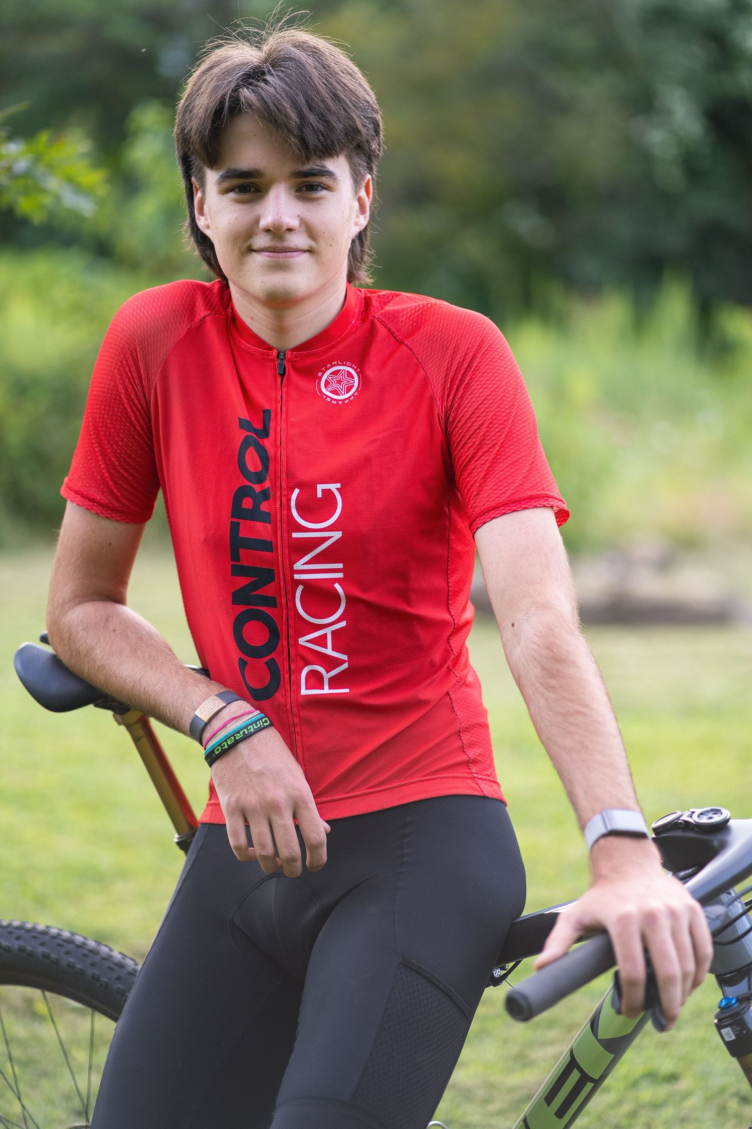 Cyclist in red jersey and black pants sits on mountain bike in outdoor setting.