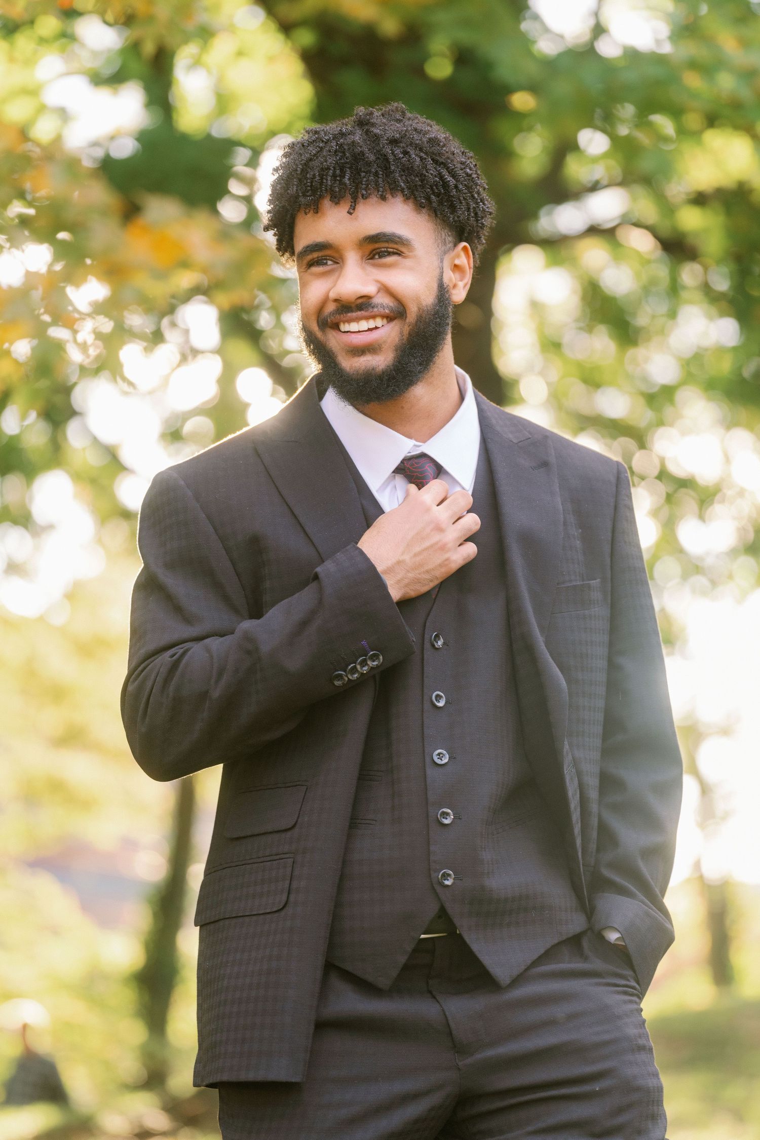 A person in a dark suit and vest poses outdoors with a cheerful expression against a blurred natural background.