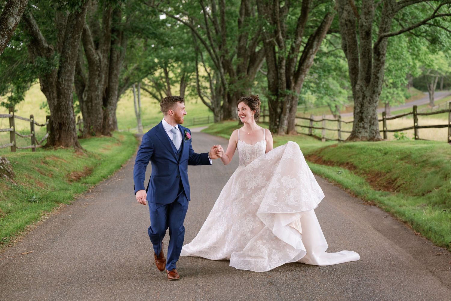 A couple in wedding attire walk and laugh together on a tree-lined country road on their special day.