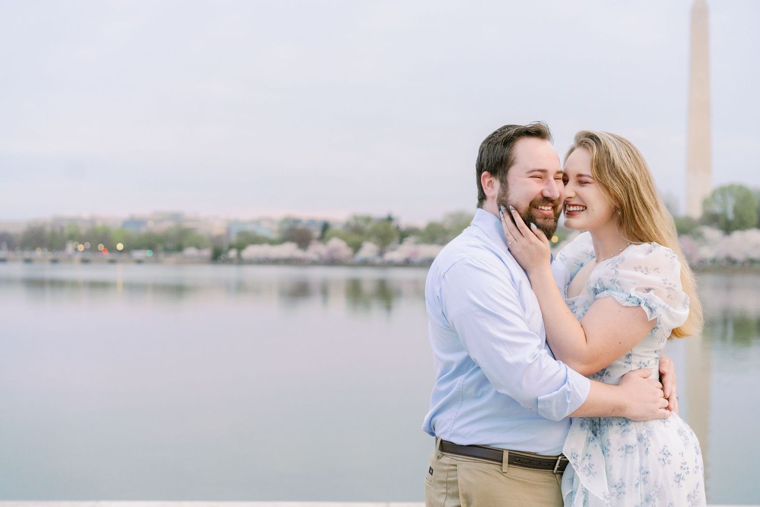 A romantic couple embraces by the Tidal Basin in Washington DC with the Washington Monument visible in the distance.